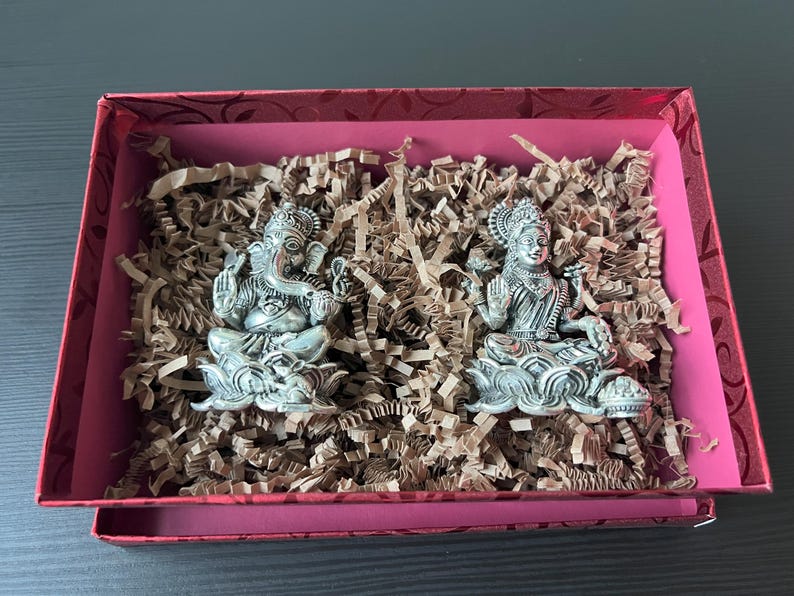 Op de afbeelding: Twee zilveren beeldjes van hindoe&iuml;stische goden, Ganesha en Lakshmi, worden getoond in een rode geschenkdoos met bruin gescheurd papier. De beeldjes zitten op lotusbloem bases.