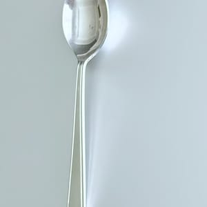 Może przedstawiać: Błyszcząca, srebrna łyżka z zaokrągloną miseczką i długim, smukłym trzonkiem. Łyżka wykonana jest z odblaskowego metalu, prawdopodobnie ze stali nierdzewnej, i umieszczona na jasnoszarym tle. Design łyżki jest prosty i elegancki.