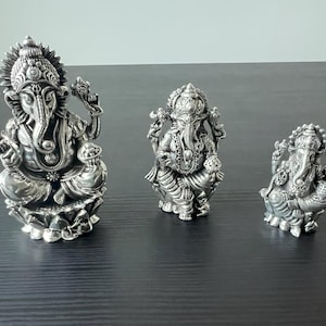 Op de afbeelding: Drie zilveren Ganesha-beeldjes in verschillende maten. Het grootste beeldje zit op een lotusbasis, terwijl de andere twee staan. Alle drie beeldjes hebben ingewikkelde details en zijn gemaakt van gepolijst zilver.