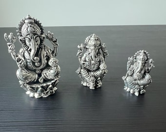 Può includere: Tre statue di Ganesha in argento di diverse dimensioni. La statua più grande è seduta su una base di loto, mentre le altre due sono in piedi. Tutte e tre le statue hanno dettagli intricati e sono realizzate in argento lucido.