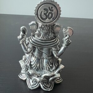 Op de afbeelding: Zilveren Ganesha-beeldje dat op een lotusbasis zit met een Om-symbool op het hoofd. Het beeldje is versierd met ingewikkelde houtsnijwerk.