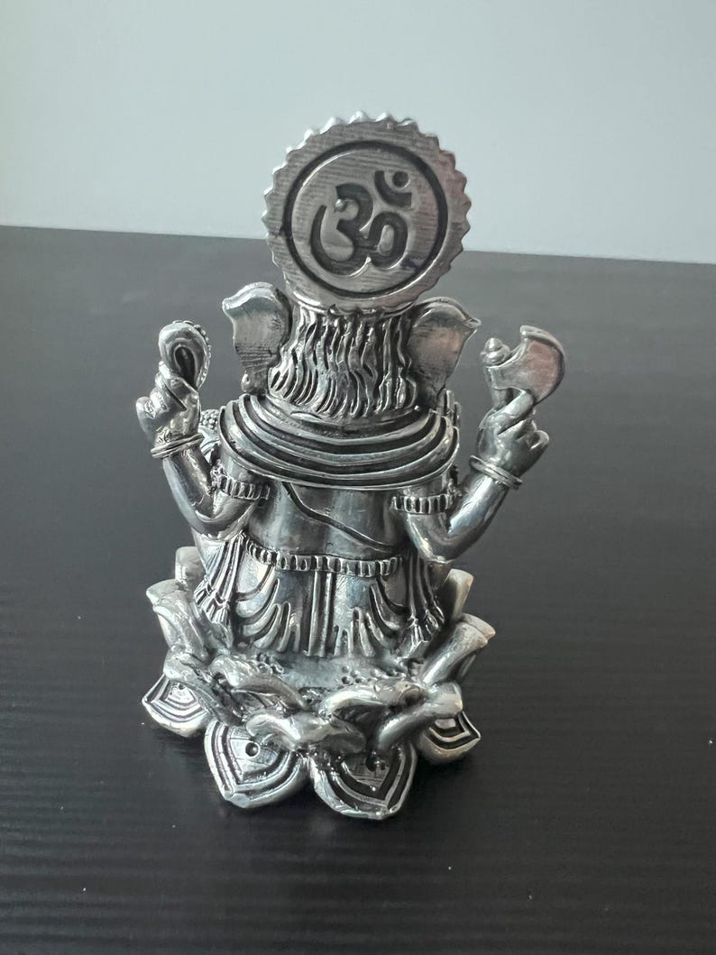 Op de afbeelding: Zilveren Ganesha-beeldje dat op een lotusbasis zit met een Om-symbool op het hoofd. Het beeldje is versierd met ingewikkelde houtsnijwerk.