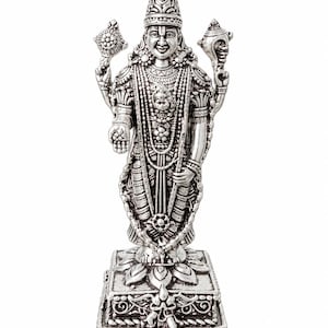 Op de afbeelding: Een zilverkleurig metalen beeldje van een godheid die op een vierkante voet staat. Het figuur draagt een versierde puntige hoofdtooi, gelaagde kettingen en armbanden. Vier armen zijn zichtbaar, met een schijf en een schelp in de handen.