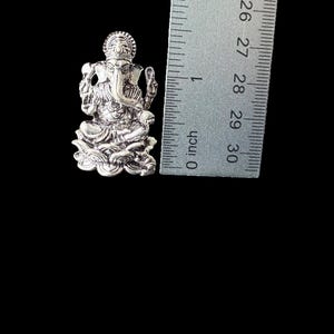 Op de afbeelding: Zilverkleurig metalen hanger met een gedetailleerde afbeelding van de hindoe-god Ganesha. De hanger is ongeveer 2,5 cm hoog en wordt naast een liniaal getoond voor schaal.