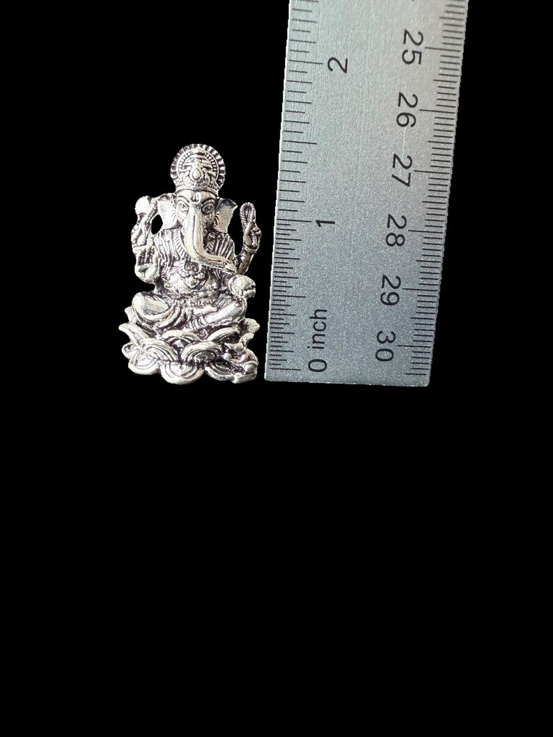 Op de afbeelding: Zilverkleurig metalen hanger met een gedetailleerde afbeelding van de hindoe-god Ganesha. De hanger is ongeveer 2,5 cm hoog en wordt naast een liniaal getoond voor schaal.