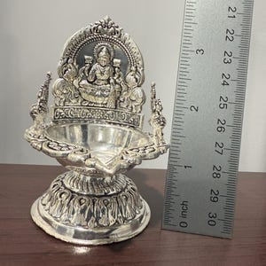Puede incluir: Diya de plata con un dise&ntilde;o detallado que presenta una figura de diosa en el centro. El diya mide aproximadamente 7,6 cm de alto y tiene una base redonda.