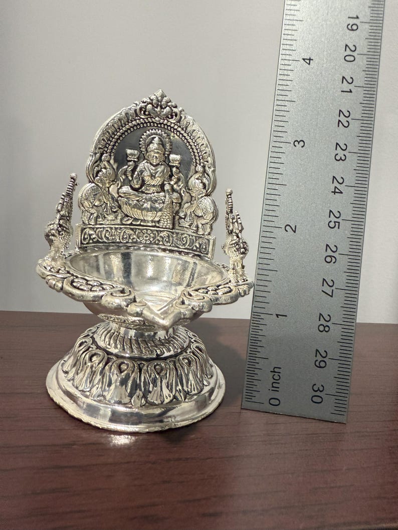 Puede incluir: Diya de plata con un dise&ntilde;o detallado que presenta una figura de diosa en el centro. El diya mide aproximadamente 7,6 cm de alto y tiene una base redonda.