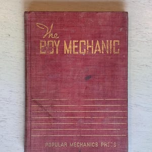 May include: A vintage book titled "The Boy Mechanic" with gold lettering on a red cover. The book is from Popular Mechanics Press. The cover has horizontal lines and some wear.