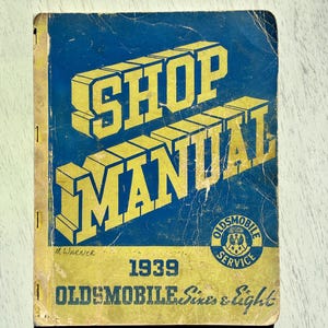May include: Vintage 1939 Oldsmobile Shop Manual. The cover features the words "SHOP MANUAL" in large yellow block letters against a blue background. The manual also includes the text "OLDSMOBILE Sixes & Eight".