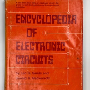 May include: An orange book titled "Encyclopedia of Electronic Circuits" with white text. The book cover includes the authors' names, Leo G. Sands and Donald R. Mackenroth, and a description of the book's contents. The book is an atlas of electronic circuit diagrams.