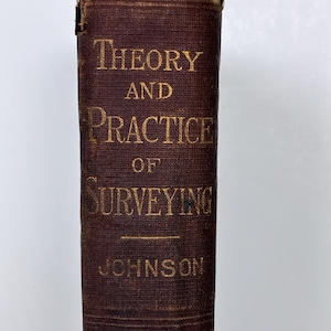 May include: A close-up of the spine of a vintage book titled "Theory and Practice of Surveying" in gold lettering on a dark brown cover. The author's name, Johnson, is also in gold. The top edge of the book shows wear.