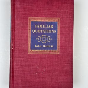 May include: A hardcover book titled "Familiar Quotations" by John Bartlett. The book has a deep red cover with a blue and gold rectangular label in the center. The label features the title and author's name in gold lettering.