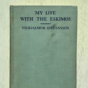 May include: A vintage book titled "MY LIFE WITH THE ESKIMOS" by Vilhjalmur Stefansson. The book has a faded teal cover with black lettering. The book is closed and in good condition.