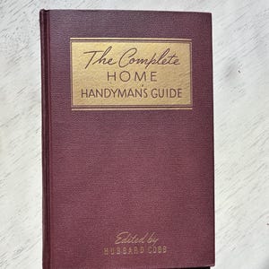 May include: A maroon-colored book titled "The Complete Home Handyman's Guide" with gold lettering. The book's cover has a textured appearance, and the author's name is printed at the bottom.