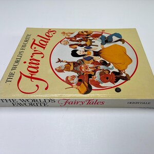 Może przedstawiać: Twarda oprawa książki zatytułowana "The World's Favorite Fairy Tales". Okładka przedstawia ilustrację Kr&oacute;lewny Śnieżki i Siedmiu Krasnoludk&oacute;w w czerwonym okręgu. Książka ma kremowe tło, a tytuł jest w ozdobnej czcionce.