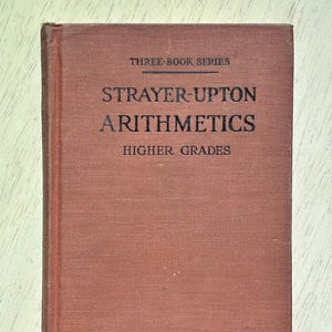 May include: A vintage textbook titled "Strayer-Upton Arithmetics Higher Grades" from the "Three Book Series." The book has a reddish-brown cover with black lettering. The book is closed and appears to be in good condition.