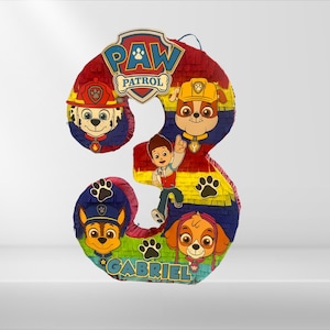 Może przedstawiać: Kolorowa piñata Paw Patrol w kształcie cyfry 3. Piñata przedstawia postacie Chase'a, Marshalla, Skye i Rydera. Imię "Gabriel" jest napisane na dole piñaty.