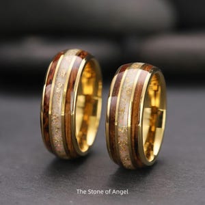 May include: Two gold rings with a wood grain and white inlay design. The rings have a gold band and a central inlay with gold flecks. The text "The Stone of Angel" is at the bottom.