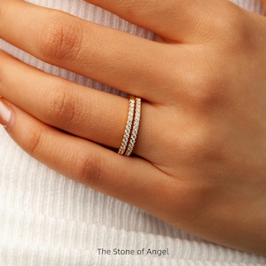 Puede incluir: Dos anillos dorados, cada uno con pequeñas piedras preciosas transparentes. Los anillos están apilados, creando un efecto de doble banda. Los anillos están en el dedo de una persona. El texto "The Stone of Angel" está en la parte inferior.
