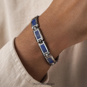 Könnte beinhalten: Ein Gliederarmband mit rechteckigen, schusswaffengrauen Metallsegmenten, die sich mit eingelegten tiefblauen Steinpaneelen abwechseln. Jedes Metallsegment enthält ein kleines pyramidenförmiges Nietendetail. Der Text TheStoneofAngel ist unten sichtbar.