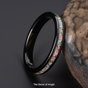 May include: A black ring with a band of colorful opal-like stones. The ring is set against a dark background and the text "The Stone of Angel" is at the bottom.