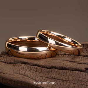 Könnte beinhalten: Zwei polierte Ringe aus Roségold mit einer glatten, reflektierenden Oberfläche. Die Ringe sind zusammen positioniert, wobei einer leicht hinter dem anderen liegt. Der Text TheStoneofAngel ist am unteren Bildrand sichtbar.