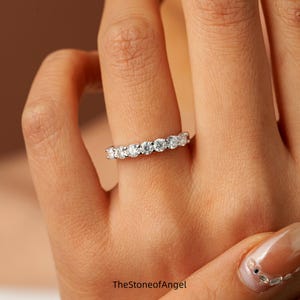 Puede incluir: Un anillo plateado con una fila de piedras preciosas redondas y transparentes. El anillo está en un dedo, con el nombre de la marca "TheStoneofAngel" visible en la parte inferior de la imagen. Un diseño clásico para cualquier ocasión.