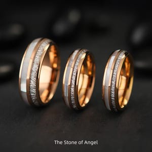 Può includere: Tre anelli in oro rosa con una fascia bianca e un intarsio argentato strutturato. Gli anelli sono esposti su una superficie scura. Il testo "The Stone of Angel" è in basso nell'immagine.