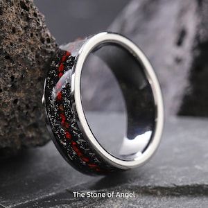 Pode incluir: Um close-up de um anel com uma faixa salpicada preta e vermelha e uma borda externa prateada. O anel tem um acabamento brilhante e polido. O texto "The Stone of Angel" está na parte inferior da imagem.