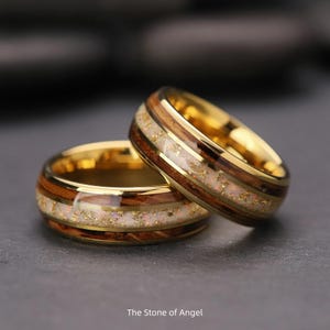 Puede incluir: Dos anillos dorados con un diseño de veta de madera e incrustaciones blancas. Los anillos tienen una banda dorada y una incrustación decorativa con motas doradas. El texto "The Stone of Angel" está en la parte inferior.