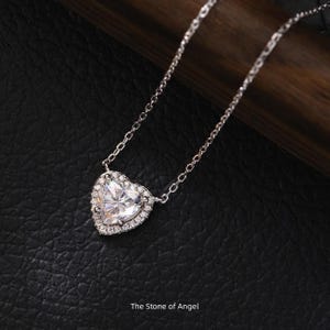 Könnte beinhalten: Eine silberne Halskette mit herzförmigem Anhänger. Der Anhänger hat einen großen, klaren Edelstein, der von kleineren Steinen umgeben ist. Die Kette ist zart und die Halskette wird auf einer dunklen Oberfläche präsentiert. Der Text "The Stone of Angel" steht unten.