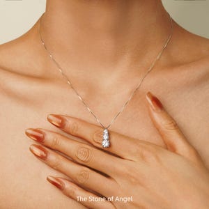 Peut inclure: Un collier en argent avec trois pierres précieuses rondes et transparentes disposées verticalement. Le collier est porté sur un cou, une main touchant le pendentif. Le texte "The Stone of Angel" est en bas.