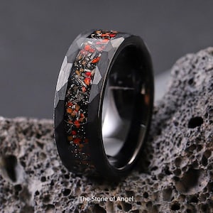 May include: Black ring with a faceted exterior and a central inlay of red and orange fragments. The ring is set against a textured gray background. The text "The Stone of Angel" is at the bottom.