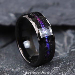 Op de afbeelding: Een zwarte ring met een facetgeslepen buitenband en een galaxy-thema inleg van paars, blauw en wit. De binnenkant van de ring heeft een sprankelend ontwerp, waardoor een hemels effect ontstaat.