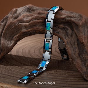 May include: A link bracelet featuring polished silver-tone metal segments alternating with rectangular blue and green iridescent opal inlays. The bracelet is draped over a piece of driftwood. The text TheStoneofAngel is printed at the bottom.