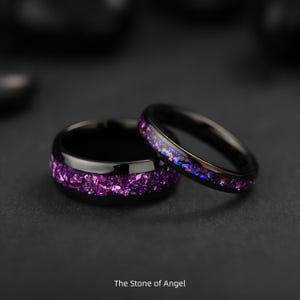 May include: Two black rings with a band of crushed purple gemstones. One ring is wider than the other. The text "The Stone of Angel" is at the bottom of the image. The rings are on a dark surface.
