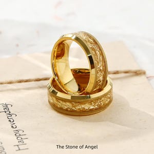 Può includere: Due anelli color oro con un intarsio strutturato marrone chiaro. Gli anelli sono impilati l'uno sull'altro, con il testo "The Stone of Angel" visibile in basso. Gli anelli sono su una superficie chiara con il testo "Happy everyday!" visibile.