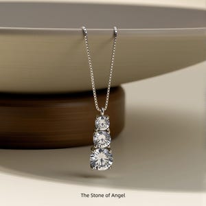 May include: A silver necklace featuring three graduated, round, clear gemstones. The pendant is suspended from a fine chain. The text "The Stone of Angel" is at the bottom of the image. The necklace is set against a neutral backdrop.