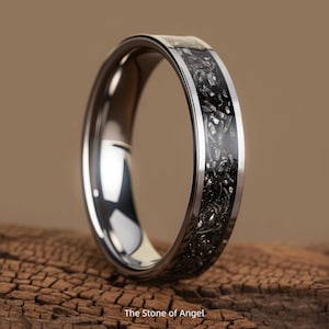 May include: A silver ring featuring a black, textured inlay. The ring has a polished finish and a silver band framing the black inlay. The text "The Stone of Angel" is visible at the bottom of the image.