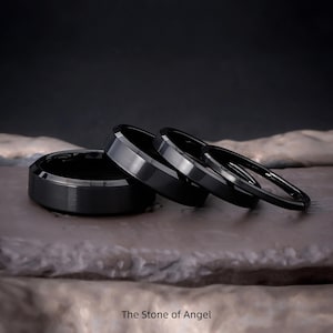 Puede incluir: Cuatro anillos negros con diferentes diseños se muestran sobre una superficie texturizada. Los anillos tienen acabados pulidos y mate, uno de ellos con una textura de metal cepillado. El texto "The Stone of Angel" está en la parte inferior.