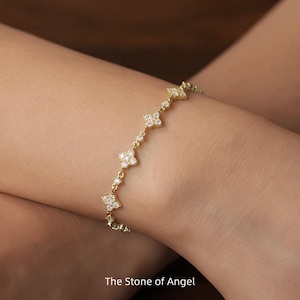 Op de afbeelding: Een goudkleurige armband met een reeks klavervormige bedels, elk versierd met kleine, sprankelende stenen. De armband is delicaat en elegant, met de tekst "The Stone of Angel" onderaan.