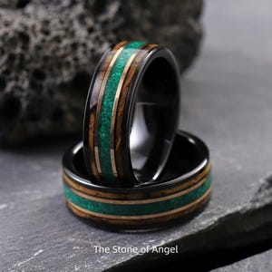 May include: Two black rings with a unique design. The rings feature a central band of green stone, framed by gold-colored metal and dark wood. The rings are stacked on top of each other. The text "The Stone of Angel" is at the bottom.