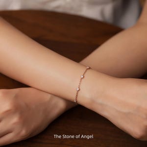 Pode incluir: Uma delicada pulseira em ouro rosa com várias pedras preciosas redondas e transparentes. A pulseira é usada no pulso. O texto "The Stone of Angel" é visível na parte inferior da imagem. O design da pulseira é simples e elegante.