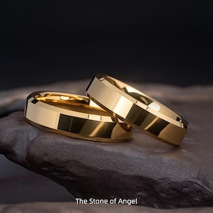 Op de afbeelding: Twee goudkleurige ringen met een facetontwerp, liggend op een donker, getextureerd oppervlak. De ringen zijn hoogglans gepolijst en reflecteren licht. De tekst "The Stone of Angel" is zichtbaar onderaan.