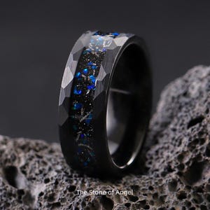 Peut inclure: Bague noire avec une surface facettée et une incrustation centrale de matériau irisé bleu et noir. La bague est posée sur un fond sombre et le texte "The Stone of Angel" est visible en bas.