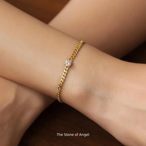 May include: A gold-colored chain bracelet with a heart-shaped, clear gemstone in the center. The bracelet is on a wrist. The text "The Stone of Angel" is at the bottom of the image.
