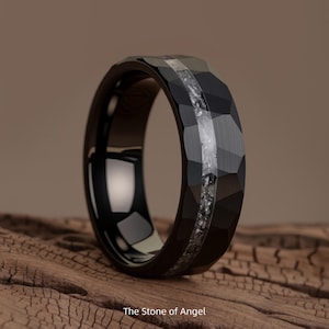 Op de afbeelding: Zwarte facetring met een centrale inleg van lichtgekleurde steen. De ring heeft een gepolijste binnenband en een getextureerd buitenoppervlak. De tekst "The Stone of Angel" staat onderaan de afbeelding.