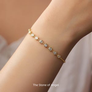 Könnte beinhalten: Ein goldenes Armband mit runden, klaren Edelsteinen. Das Armband wird am Handgelenk getragen und weist eine Reihe von Steinen in Zargenfassung auf. Der Text "The Stone of Angel" befindet sich am unteren Rand des Bildes.