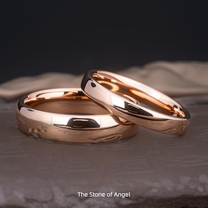 May include: Two rose gold wedding rings with a delicate engraved design. The rings are presented on a neutral surface, with the text "The Stone of Angel" at the bottom. The bands have a polished finish and a classic style.
