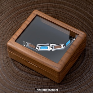 Puede incluir: Una caja de presentación rectangular de madera con tapa de cristal transparente que contiene una pulsera de eslabones. La pulsera presenta eslabones de metal plateado que alternan con segmentos de incrustaciones azul brillante.